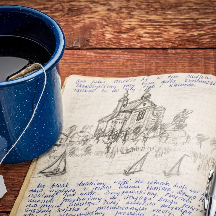 journal with handwriting and drawing in pencil in a notebook against rustic picnic table with cup of tea
