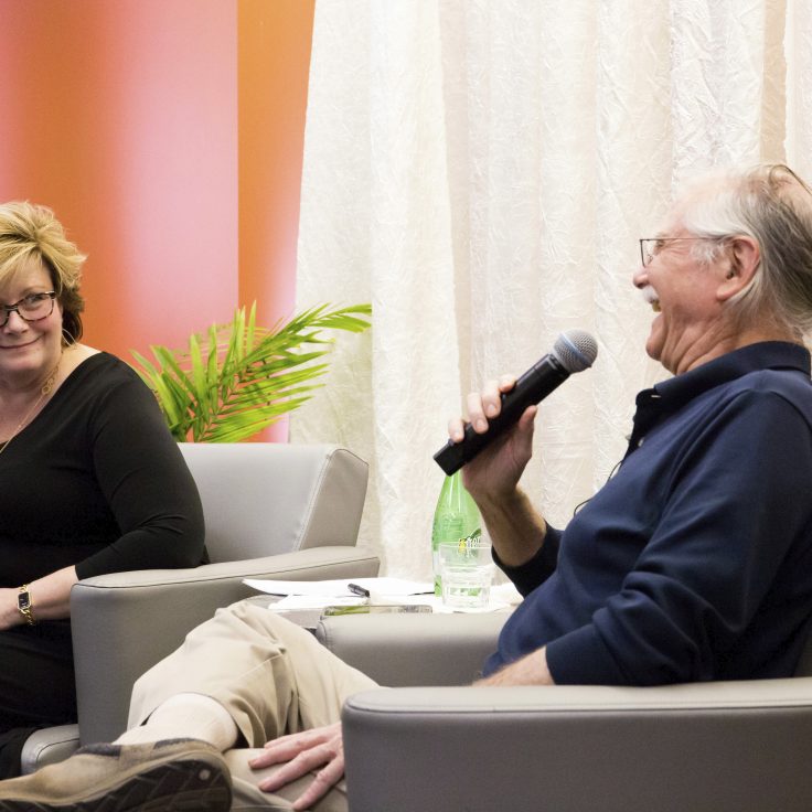 an older man laughs into a microphone, sitting in a comfy chair, while a blonde woman looks on with interest