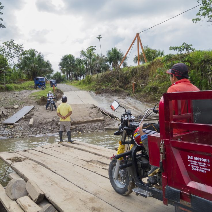 red rickshaw on rugged barge crosses river as people on opposite shore await