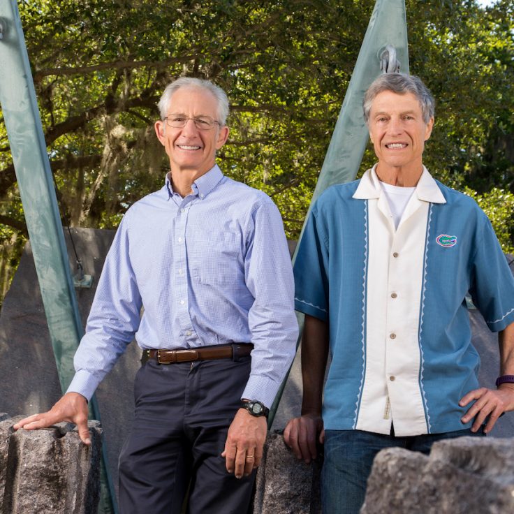 two smiling men sit on boulders with teal industrial structure behind them