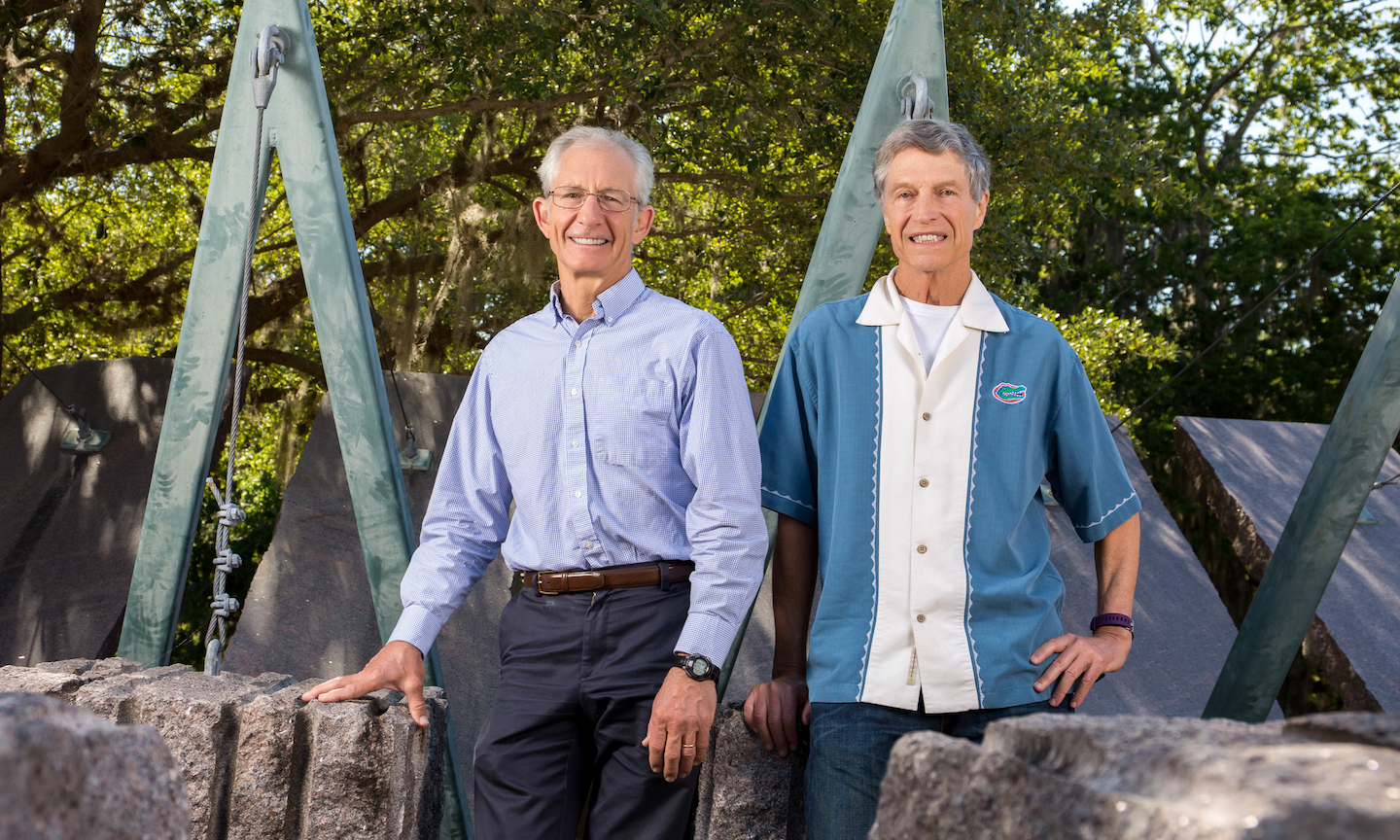 two smiling men sit on boulders with teal industrial structure behind them