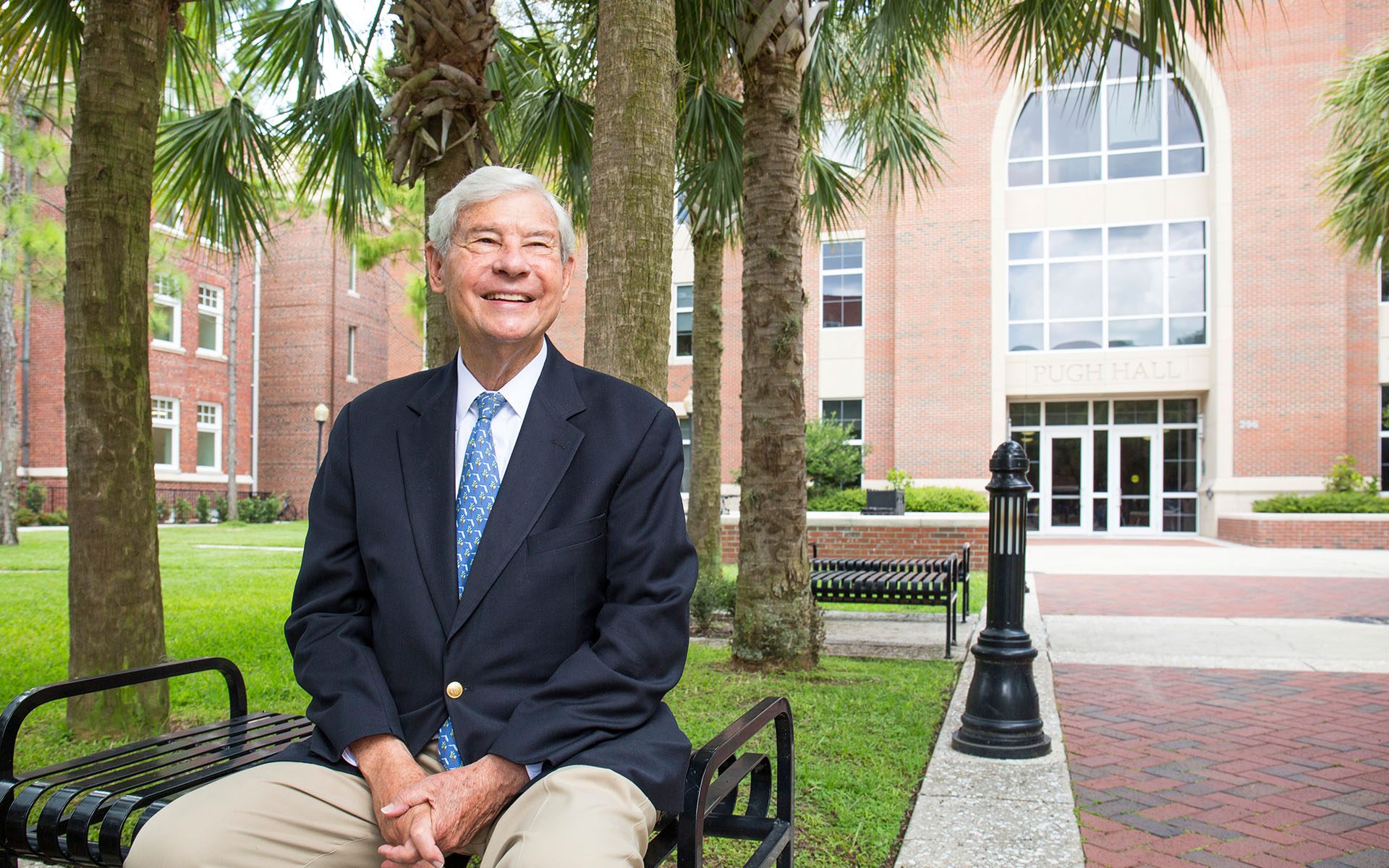 smiling grandfatherly man in suit sits on bench outside statuesque collegiate building