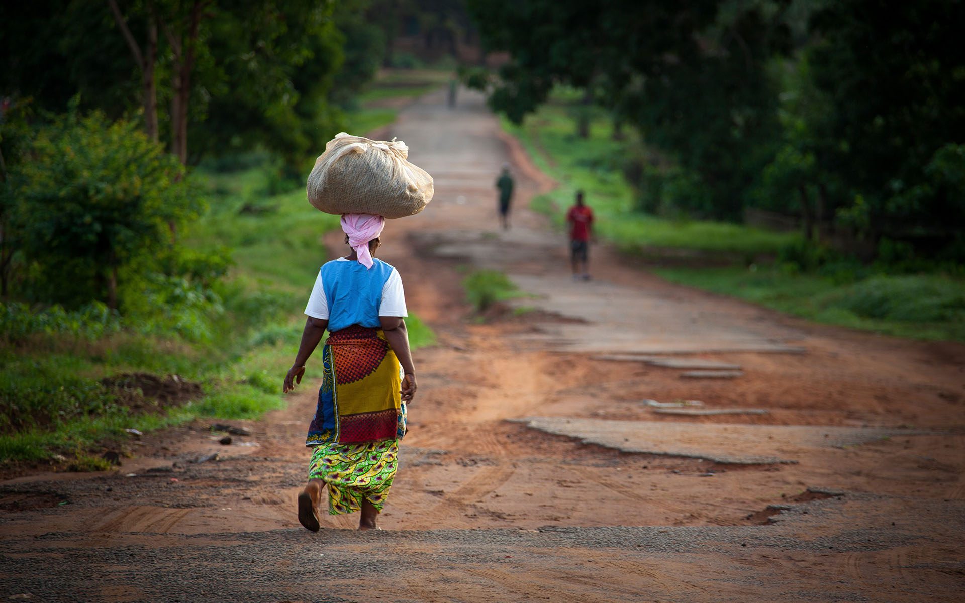 woman in traditional African garb walks down dirt road, a parcel balanced upon her head