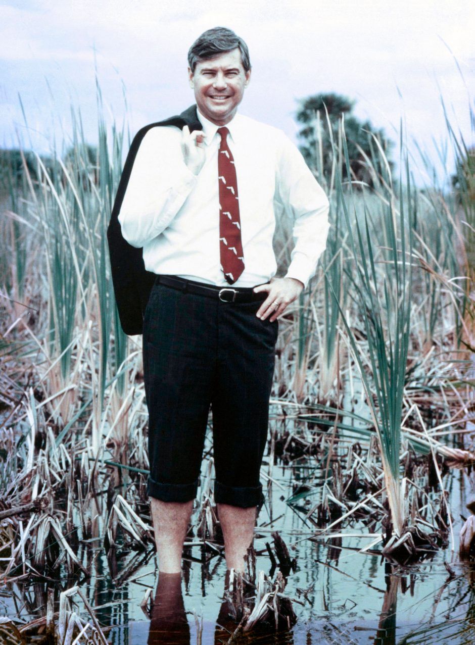 well-dressed man stands in calf-deep in swamp with jacket over shoulder and pants rolled up