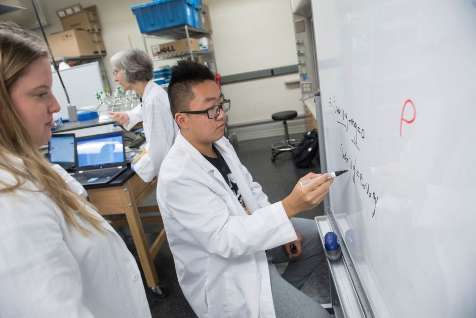 young male student in white coat writes equations on whiteboard