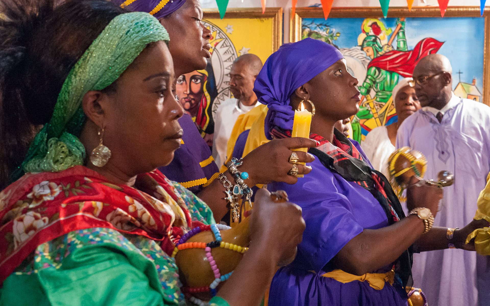 Haitian women adorned in vivid colors hold candles and instruments