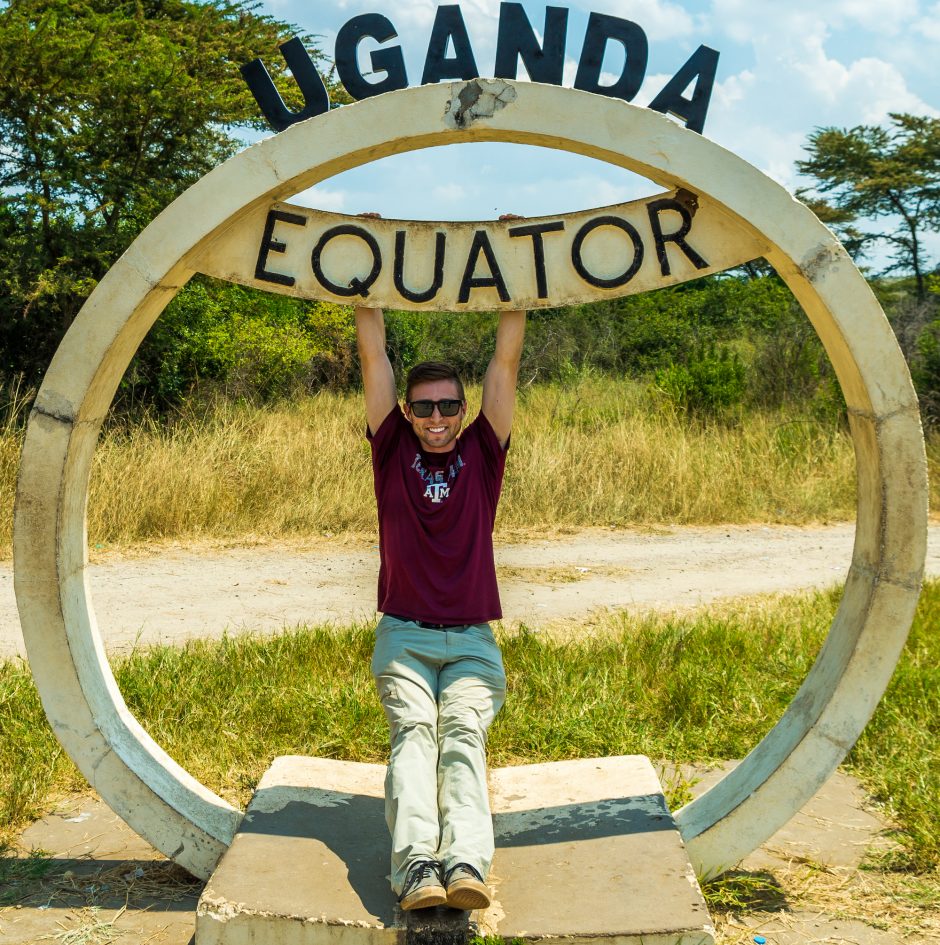 young man beams as he hangs from a round structure with a sign reading "Uganda" and "Equator"