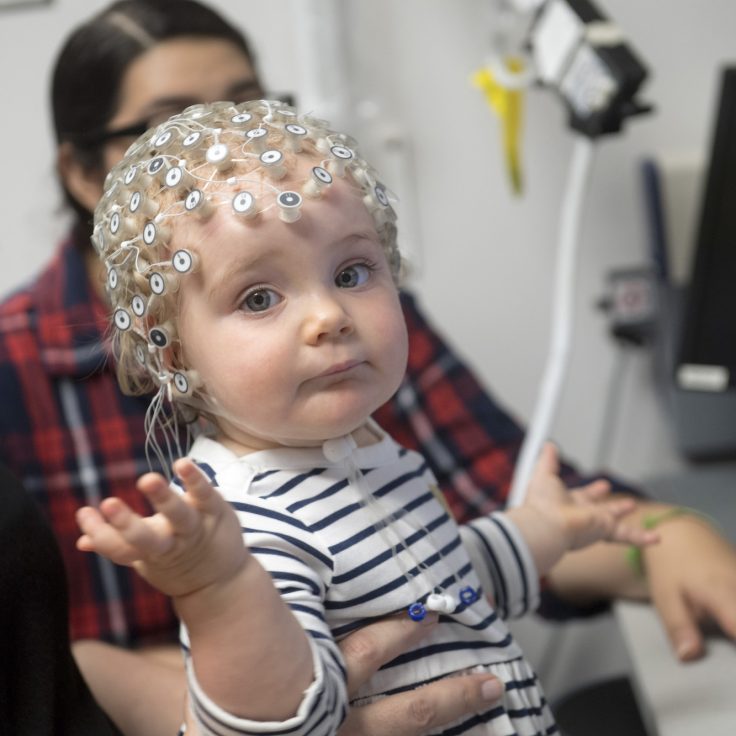 infant wearing an EEG cap waves her hand toward the camera
