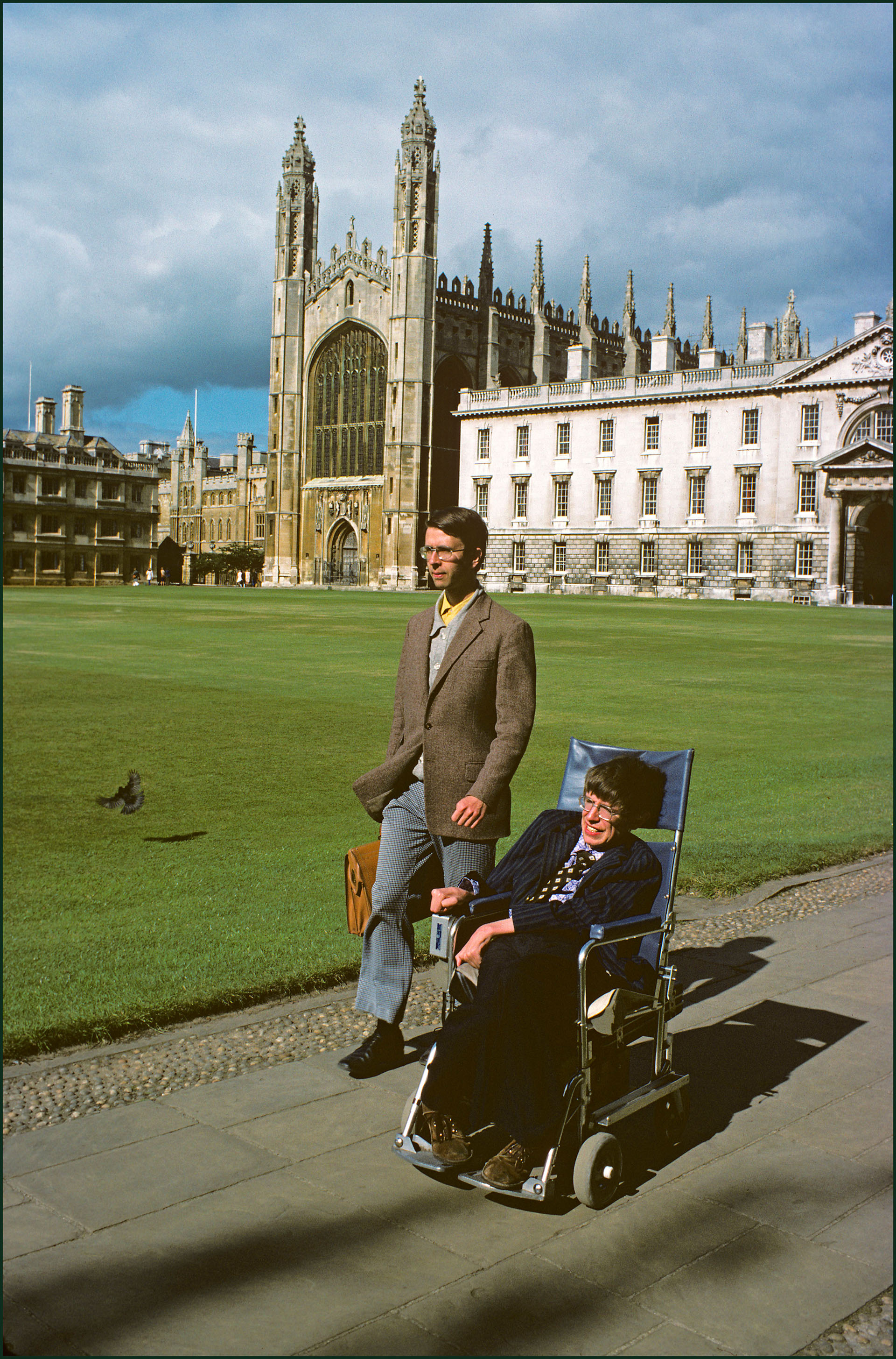 Hawking in chair with colleague alongside and majestic collegiate building in background