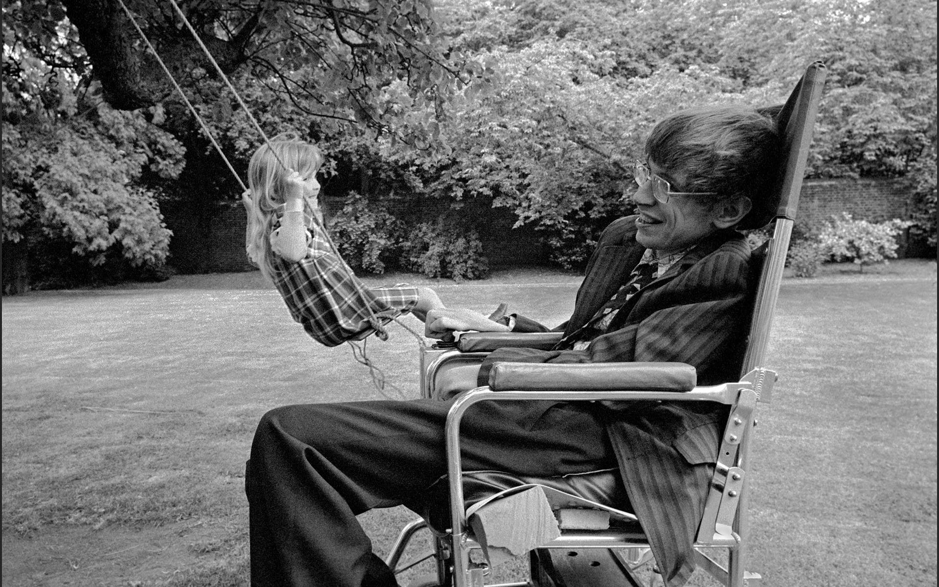 Stephen Hawking in chair as young girl swings on tree swing in background