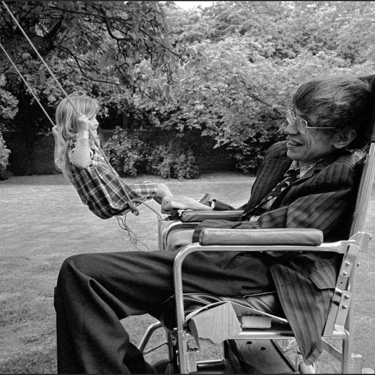 Stephen Hawking in chair as young girl swings on tree swing in background