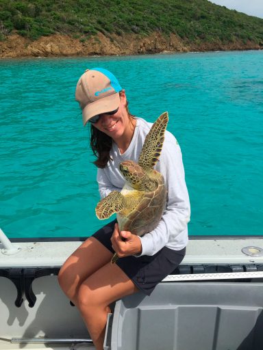 young woman sits on edge of boat holding green turtle with its flippers in the air like it just don't care