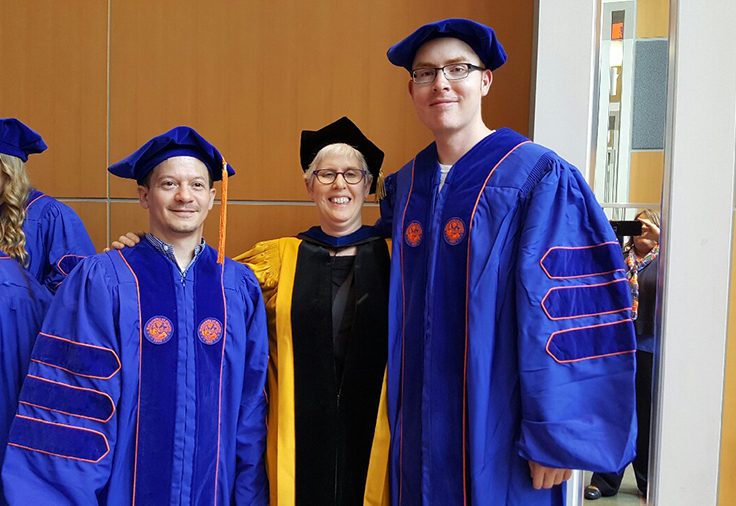 Dr. Shifra Armon stands between two graduating students, all wearing their respective ceremony attire