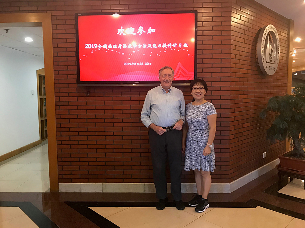 Francisco Marcos Marin and Xuhua Lucia Liang stand in front of a brick wall that has a TV display showing a red background with the text, 2019, and Chinese characters displayed in white