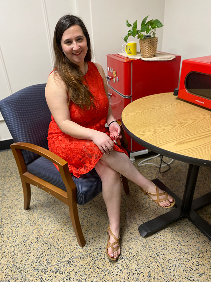 Dr. Gabriela Puscama, wearing a red dress, is seated at a table with a red appliance placed on its surface. There is a red mini-fridge in the background.