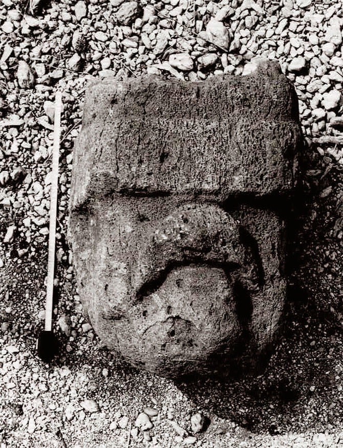 A photograph of the lost head, eroded due to time and elements. A tape measure demonstrating its size lays next to it.
