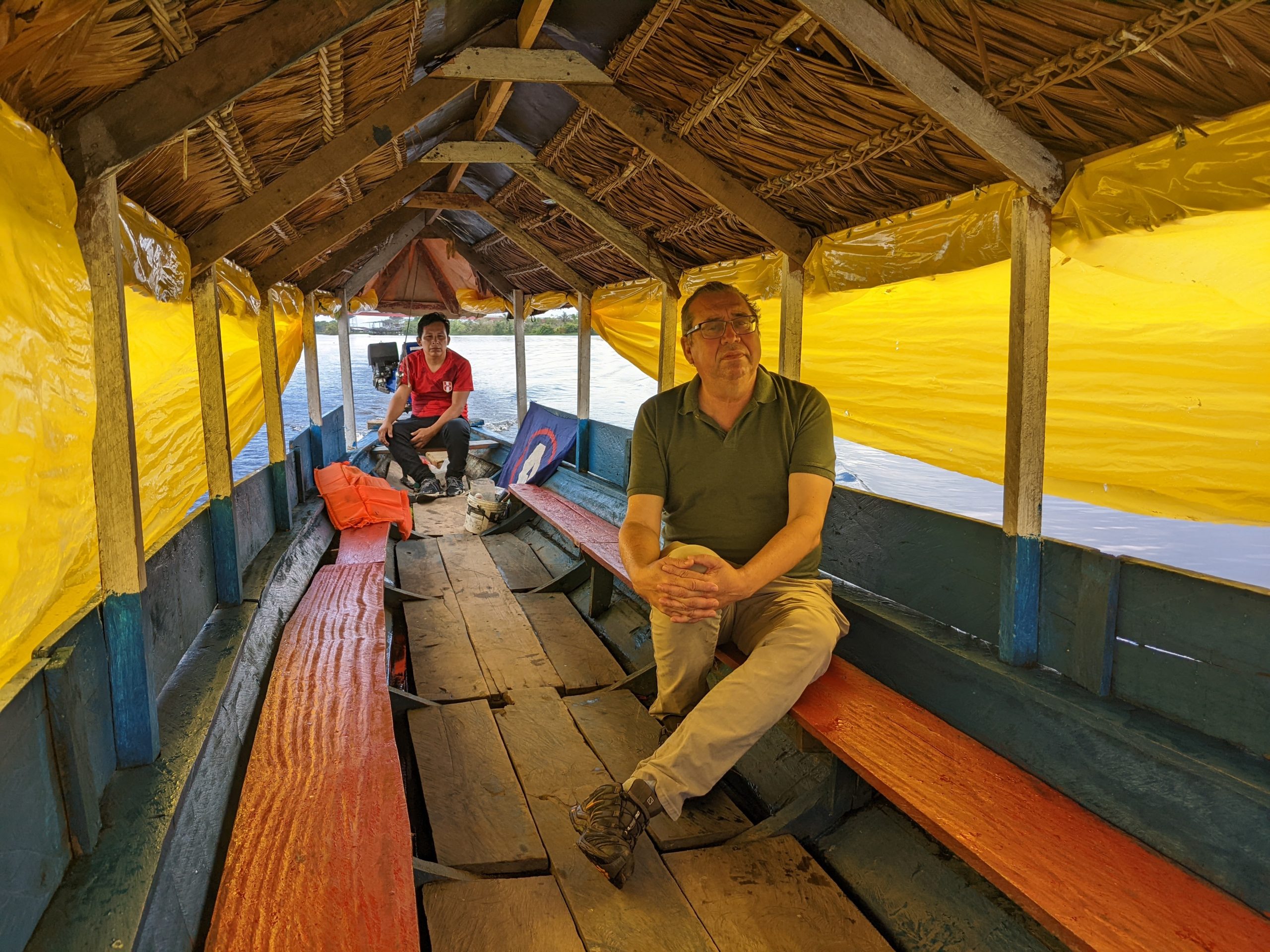 The interior of long, narrow boat with two passengers: Oyuela-Caycedo and a driver. It has two benches on each side, yellow tarp walls, and a wooden roof.
