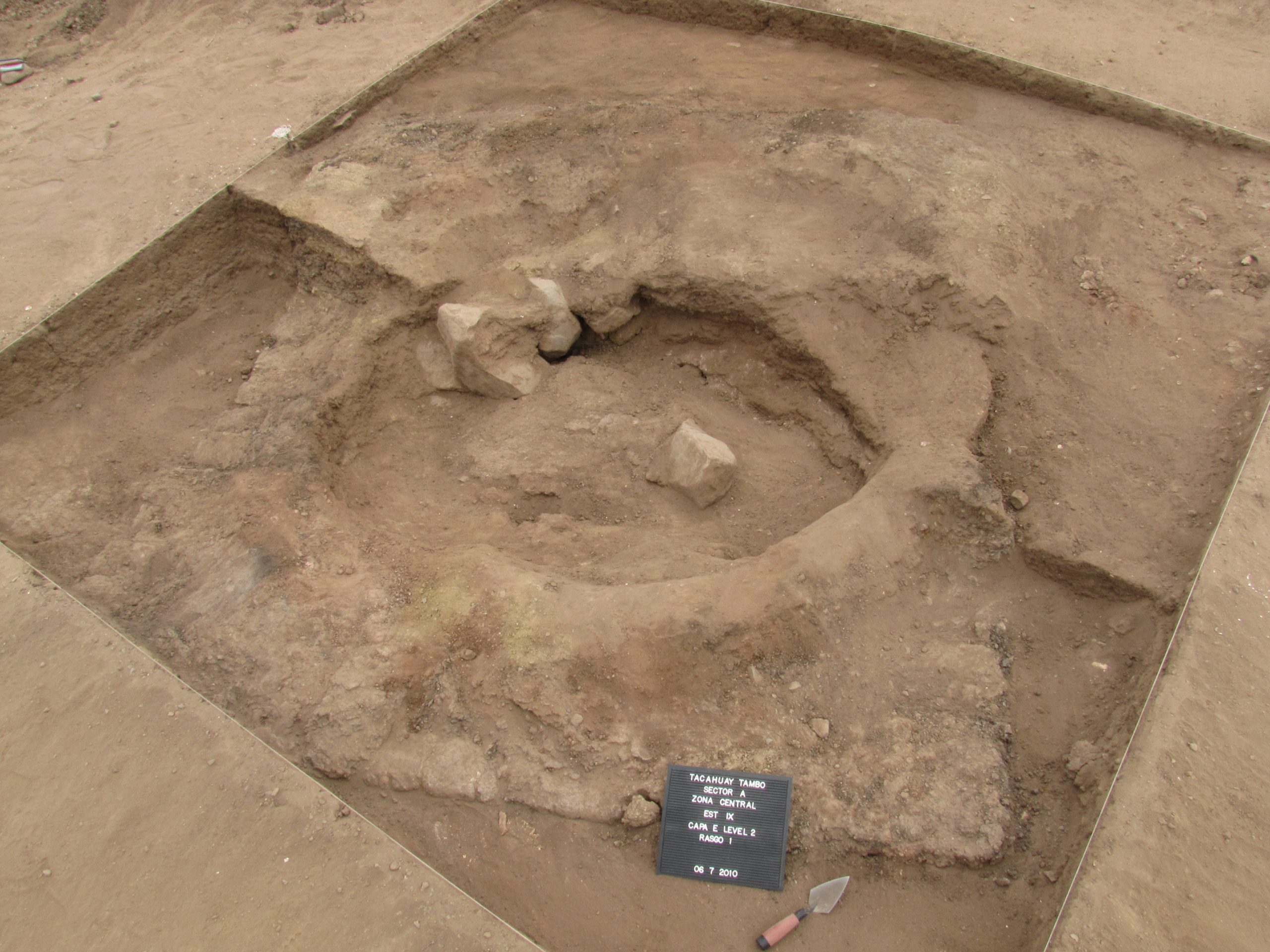 A photo of the square-shaped excavation site of the Tacahuay Tambo tomb. All that is visible so far is a mostly filled-in, circular hole carved into a square rock. 