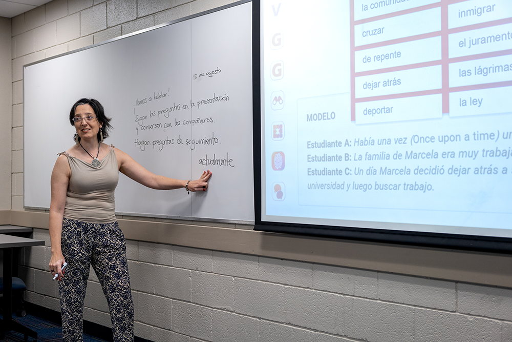 María Victoria Muñoz pointing to a whiteboard, next to a PowerPoint presentation. 