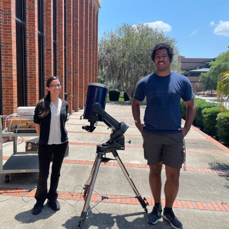 two students stand near a telescope outside of an academic building