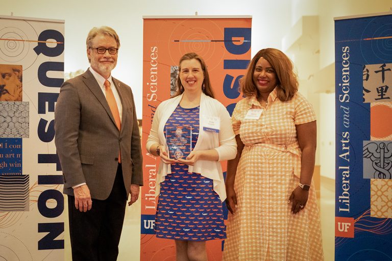 A professional man and a woman stand with a woman in the middle holding a plaque. UF banners celebrate the occasion behind them. 