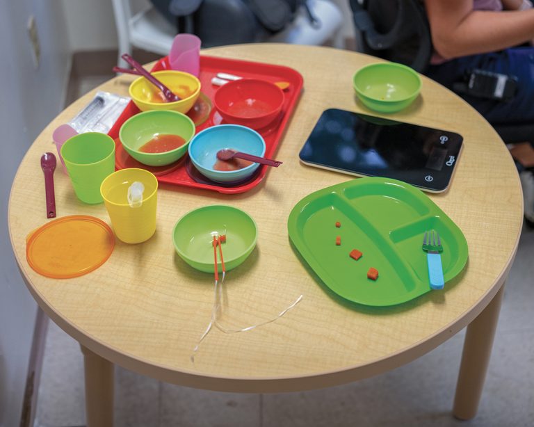 plastic bowls and cups fill a table during a feeding session, showing the soft foods, drinks, and specialized tools used for feeding