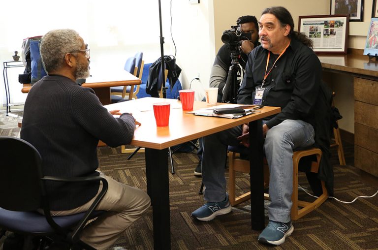 A man sits across from an interviewee, along with a camera crew, to film an interview.