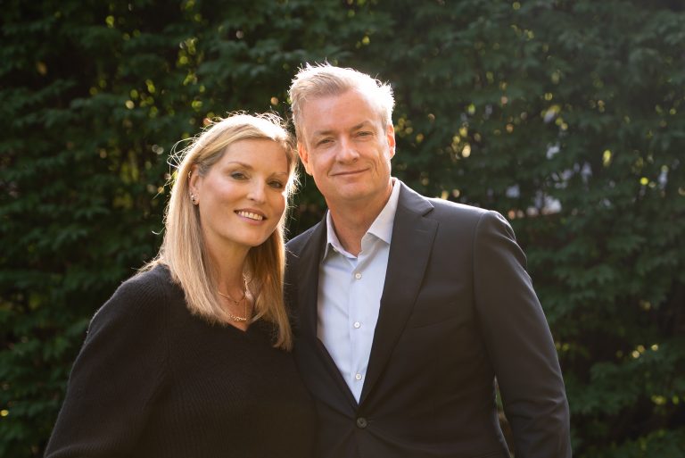 A man and a woman dressed professionally stand in front of a wooded landscape.