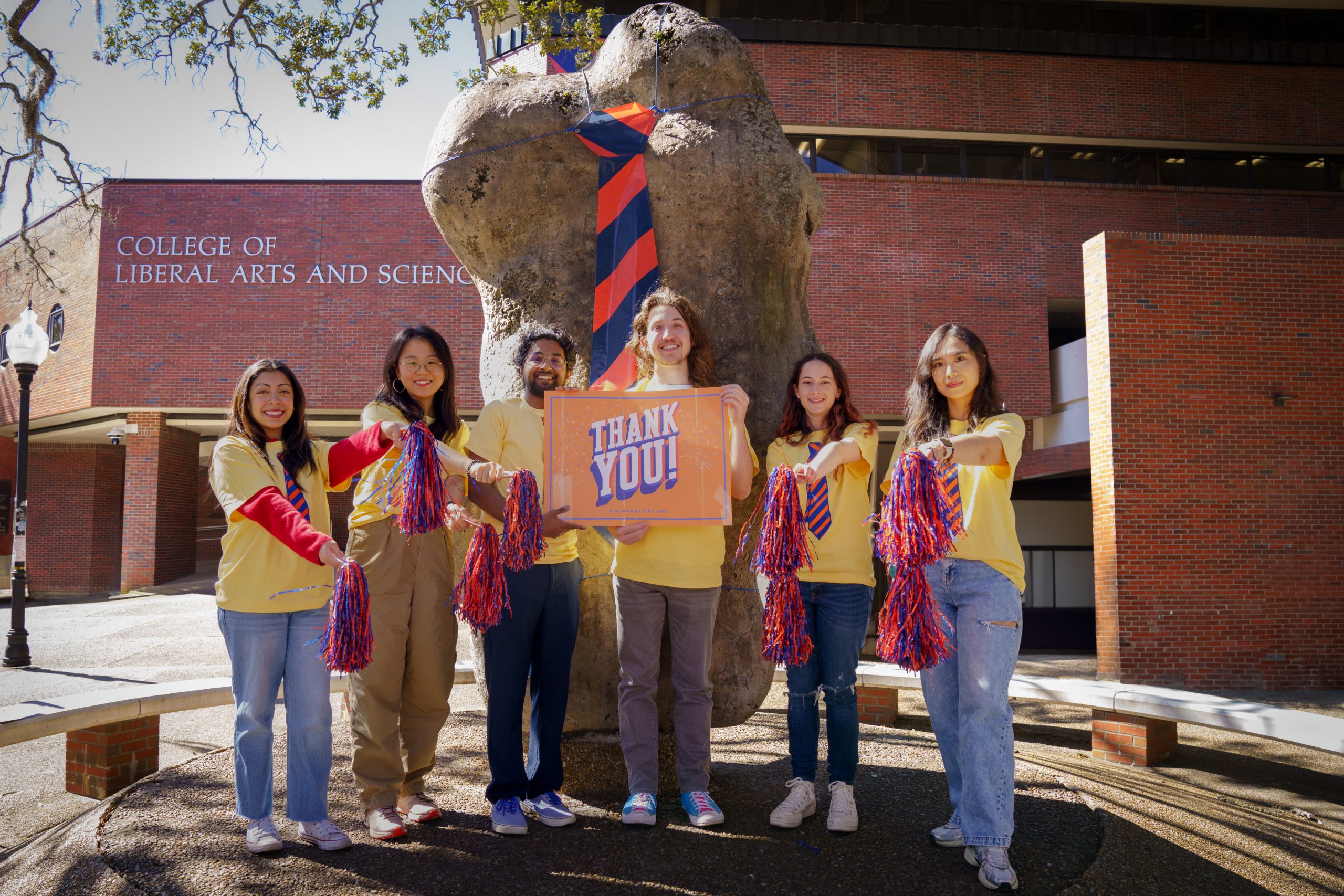 Five students dressed in yellow and the iconic Two Bits tie hold a "Thank You" sign in front of the Turlington rock.