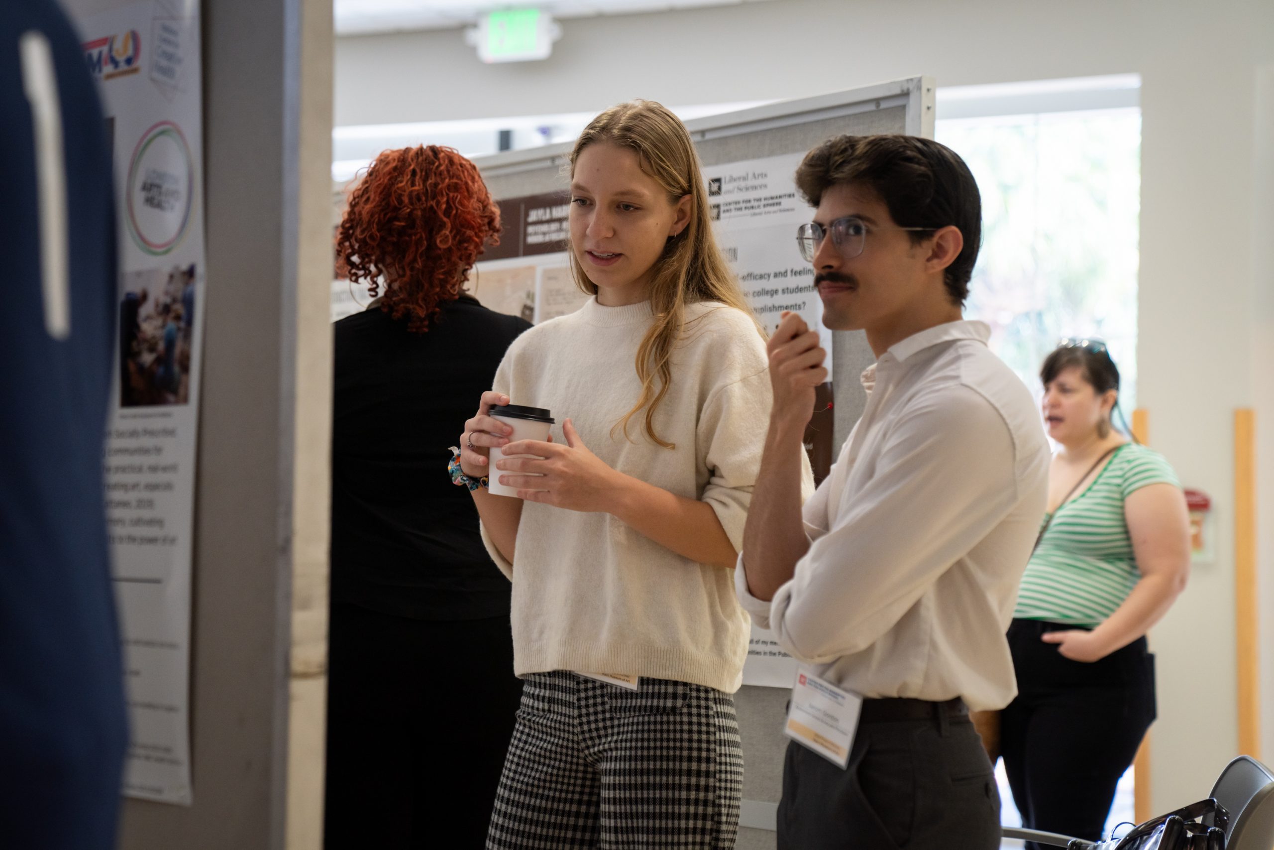 Jace Digby, holding a cup of coffee, presents her board to a fellow student. 